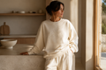 Woman wearing a cream-colored sweatshirt with 'CMC' logo in a minimalistic room.